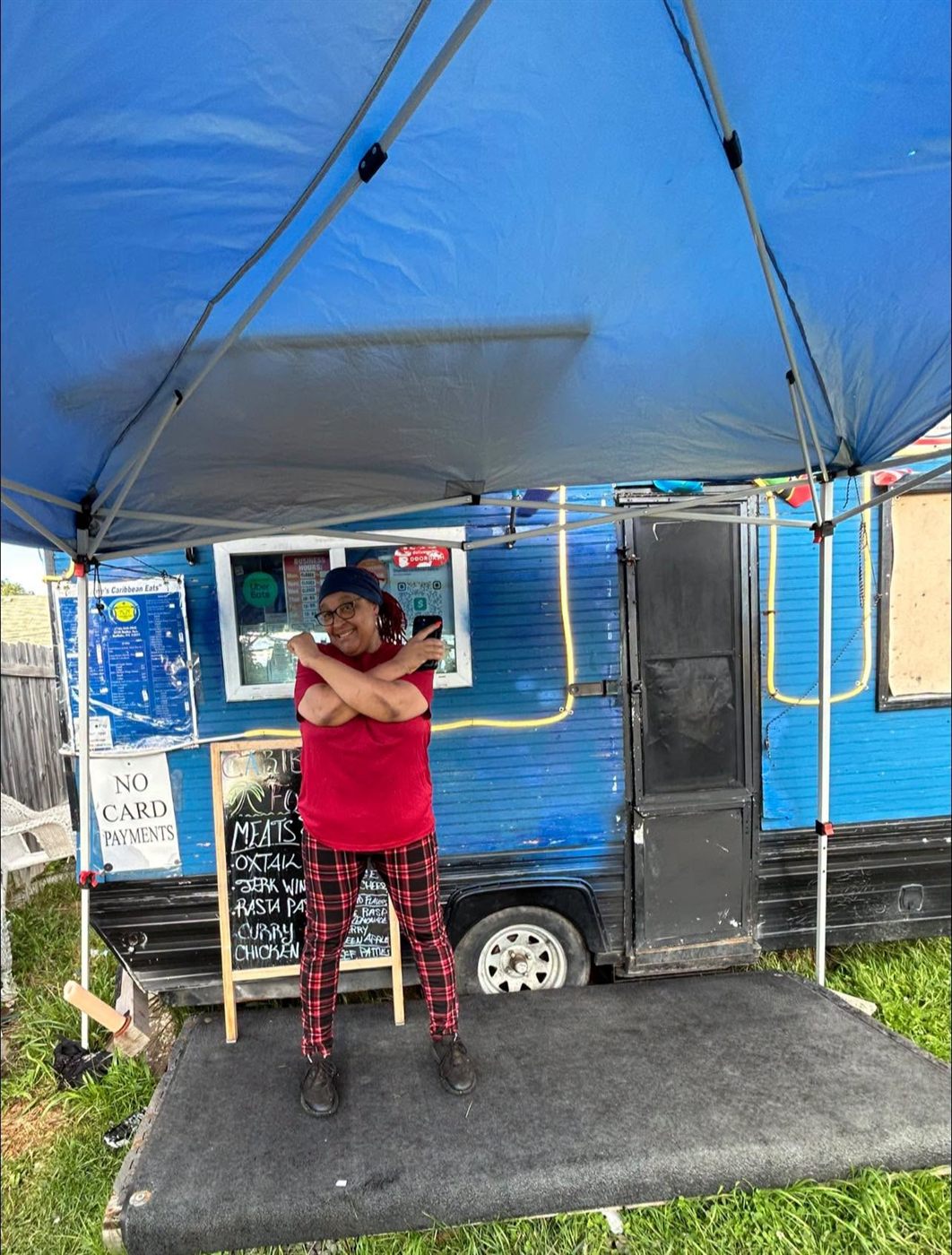 Sherrie standing at her blue Caribbean food truck in Buffalo, the day's menu chalked on a board beside her